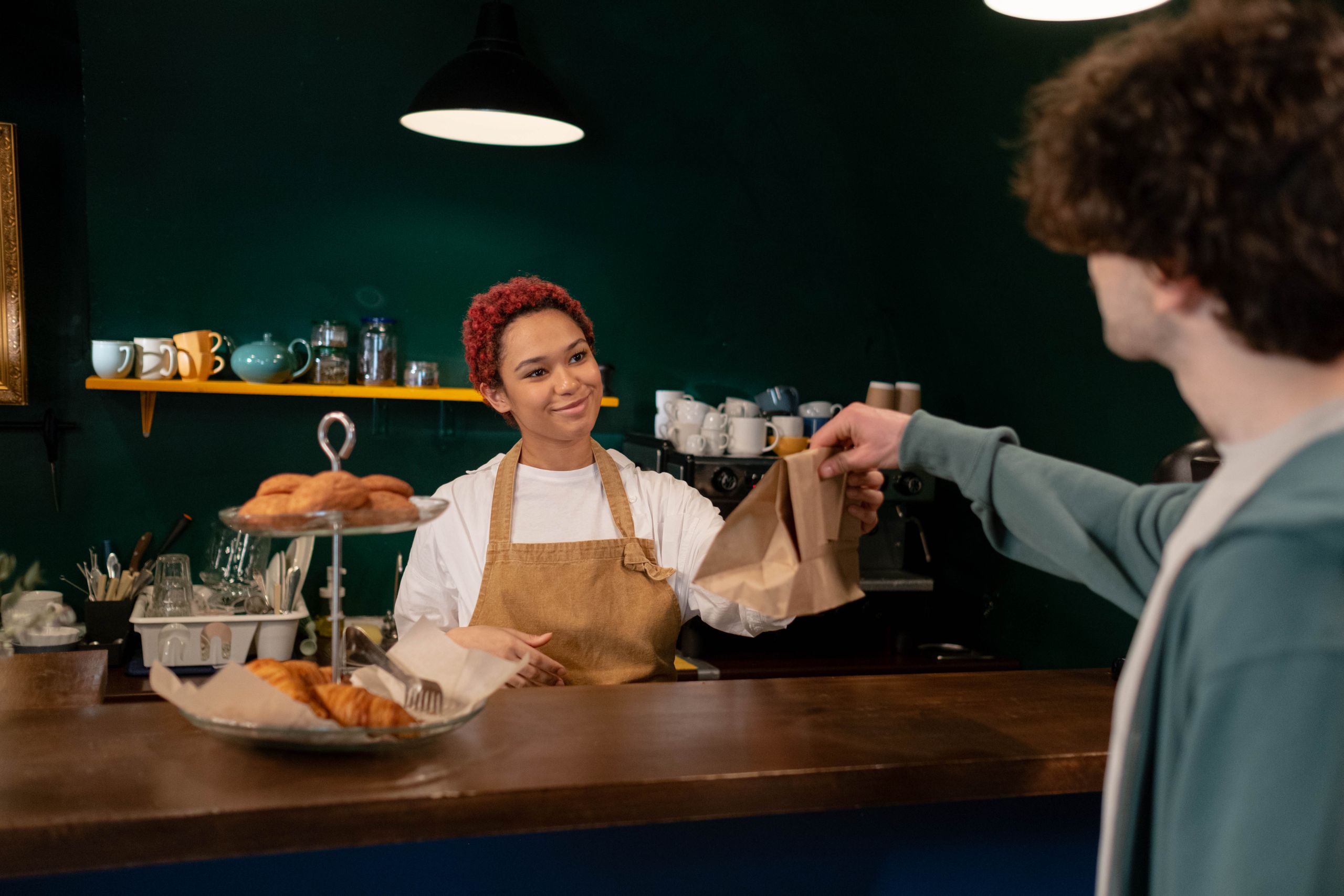 A friendly barista hands over pastries to a customer at a cozy café counter. Warm ambiance indoors.